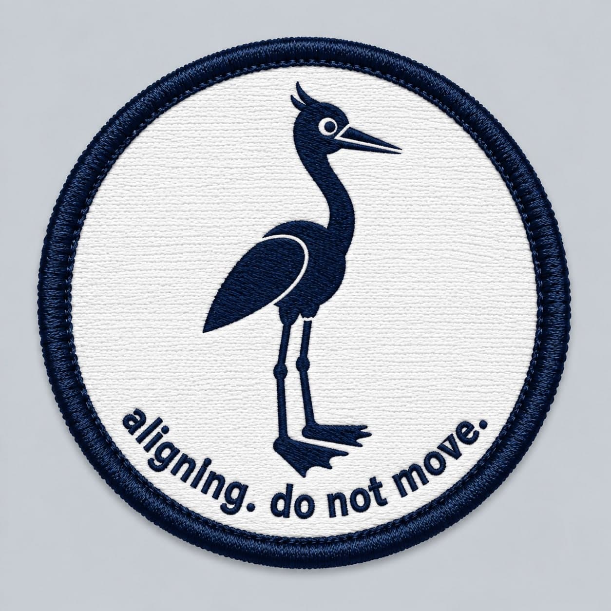 Round white embroidered patch with a navy heron standing still above the words aligning. do not move.